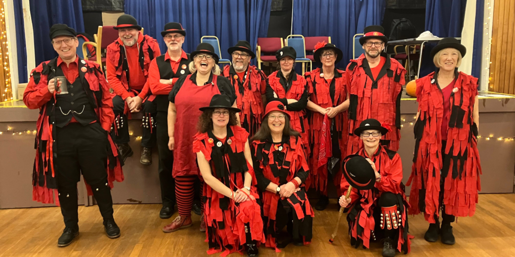 Members of Red Cuthbert Morris dressed in red and black and wearing rag coats, trying to smile for the camera. Some look happy, some grim, and some magical!